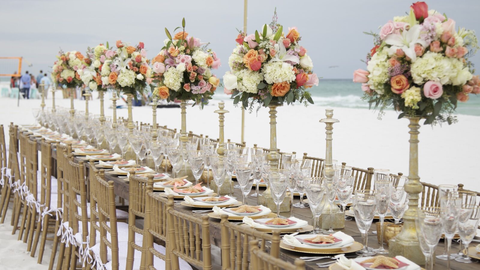 Long beach reception tables with tall peach and white floral centerpieces on the sand