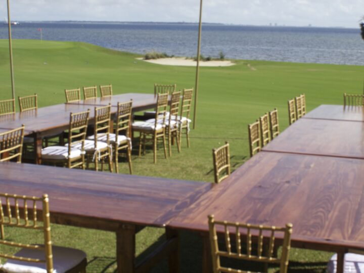 Wooden farm tables with gold Chiavari chairs on a waterfront lawn